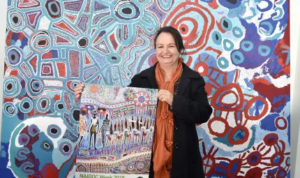 Cheryl Moggs, wearing a black jacket and orange scarf, holding the 2018 NAIDOC week poster ‘tarumunggie – woman.’ She is standing in front of a large, colourful canvas artwork.