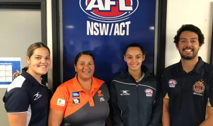 Three women and one man wearing AFL polo shirts stand in front of a large sign which says AFL NSW/ACT.