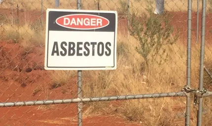 A sign reading “Danger Asbestos” attached to a gate with a background of red dirt and dry grass.