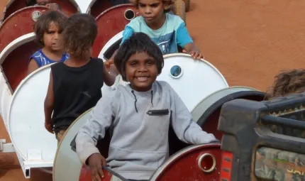 Young Aboriginal children sit in and stand next to a series of barrels mounted on wheels.