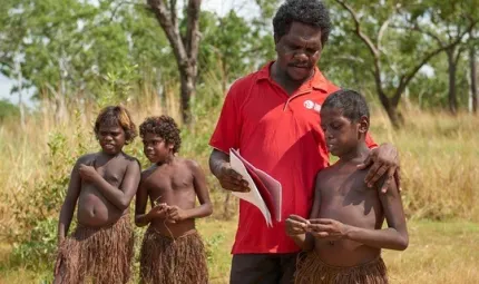 Two Aboriginal boys in grass skirts stand at left and behind an Aboriginal man in a red shirt and boy in a grass skirt in the foreground. The man holds some papers on which he and the third boy are focussed. In the background is long grass and trees.