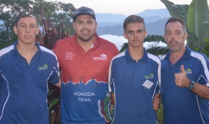 Two adult males and two young men dressed in mainly blue shirts, stand arm in arm in front of tropical foliage atop a mountain. In the background is mountain terrain and low-lying cloud.