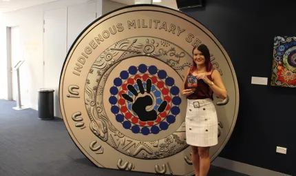 A woman in pale skirt and dark top stands next to a large replica of a coin, which features a hand in the middle of concentric circles, made up of black, red and blue dots. The coin is gold in colour. In the background are walls and a carpeted floor.