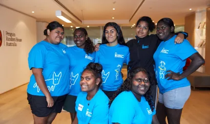 Seven young Aboriginal women, most wearing blue t-shirts stand in a group in a room with a board floor and a book case in the background. At left on the wall are the words Penguin Random House Australia.