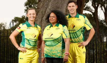 Three Indigenous people dressed in yellow and green cricketing uniforms featuring Indigenous design patterns. In the background is a tree and black iron fence.