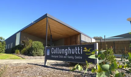 Large sign in foreground with the words Cullunghutti Aboriginal Children and Family Centre. In the background is a building with shrubs and around it. Behind the building is a sail and a clear blue sky.