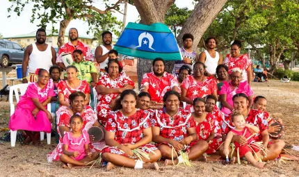 Group of people ranging from elderly to young children sit on the ground or seats or stand in front of a tree. On the tree is a blue, white and green flag. In the background are more trees, a car, a house and more people.