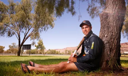 Young Aboriginal man in dark blue hat, shirt and shorts sits under a tree leaning up against the trunk while holding a cricket bat. In the background is a grassed area, a sign, many trees and a large brown hill.