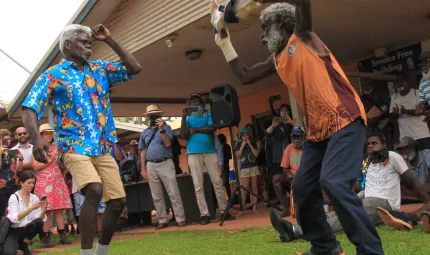 Two men dancing in front of a crowd. The dancers are wearing colourful tops and dancing with material in their hands. They are dancing in front of a crowd of Indigenous and non-Indigenous peoples