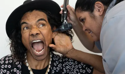 An Indigenous man in hat and black and white shirt opens his mouth and holds his ear as an Indigenous nurse applies an instrument to his ear.