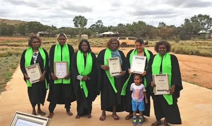 Six Aboriginal women dressed in black with bright green sashes draped around their necks and one Aboriginal child stand on a flat surface with grass and trees, a car and buildings in the background. Most of them hold a framed certificate in their hands.