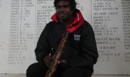 Elijah Douglas, holding his didgeridoo, sits in front of a wall of remembrance.