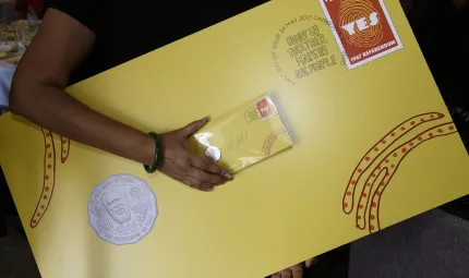 Indigenous person holding a large yellow envelope with ‘Yes’ stamp and new Eddie Mabo coin attached along with a regular size envelope of the same design. The envelope includes some Indigenous design.