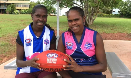 Two young Aboriginal women sit at a picnic table, holding an AFL football in their hands. One wears a blue and pink football jumper, the other wears a blue and white striped football jumper. In the background is a grassed area, trees and buildings.