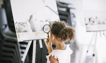 A young girl with her brown hair drawn back holds a magnifying glass up to a photo of people next to a building.