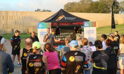 Group of people with backs to us dressed in running wear look at man in grey shirt holding microphone standing in front of a small tent with banners either side saying Closing the Gap and on the tent are the words Run Sweat Inspired.