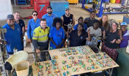 A group of people in a shed stand behind a table covered with cigarette lighters and toothbrushes. Under the table is equipment and in the background are steel racks, cupboards and machinery.