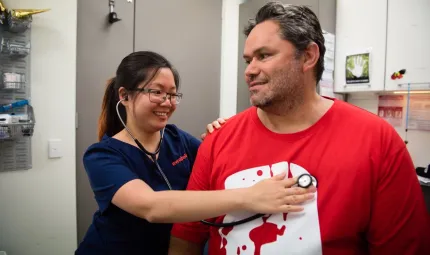 A woman in blue uniform holds a stethoscope to a man’s chest. He wears a red and white t-shirt. In the background are cupboards and racks of medical equipment.