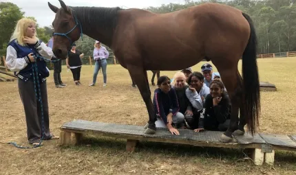 Young girls and two police officers squat behind a horse but are seen through the gap between the front and rear legs. At the head of the horse is a young woman holding its reins. In the background are more people, a grassy area and trees.