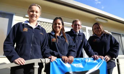 Four young adults wearing blue jumpers standing in front of a building and hanging onto a rail.