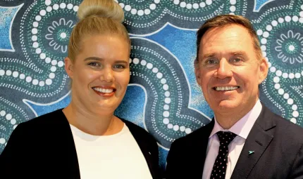 Indigenous woman in white top and black jacket with non-Indigenous man dressed in suit and tie stand in front of wall covered with blue, white and green Indigenous designs