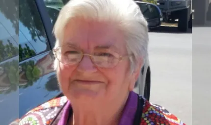 An elderly Aboriginal woman in a colourful polo shirt standing outside next to a car.