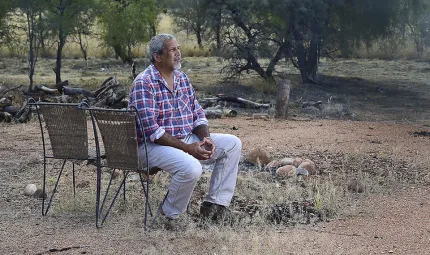 A man in checked shirt and jeans sits on a chair next to a campfire. In the background are trees and logs.