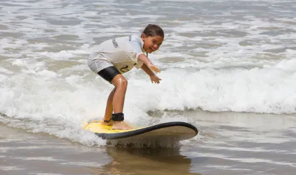 Young Aboriginal child in black shorts and white shirt stands on a surfboard riding a half-metre high wave.