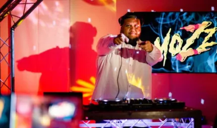 A young man stands behind disc jockey mixing desk. In the background is his shadow, a TV screen and red and yellow lights on a wall. He points to camera.