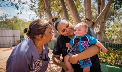 Two Aboriginal women smile at an Aboriginal child held by one of them. The child wears a blue jumpsuit with Aboriginal designs on the front. In the background is a flower bed, two fences and trees.