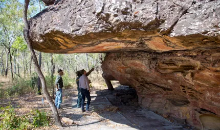 Laura Rangers stand under an overhanging rock with Aboriginal art painted on the rock walls and roof. In the background are trees and grass.