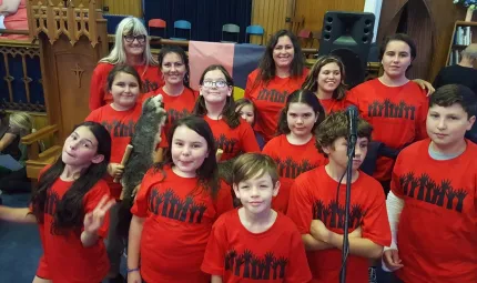 Two adult women and twelve children stand in a room behind a microphone, all wear red t-shirts with a graphic of raised hands.