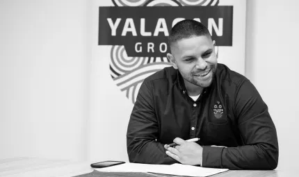 Young adult man in dark shirt sits at a table. In the background is a logo including the words Yalagan Group.