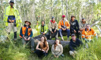 A group of people sit on or in front of a fallen tree and face the camera. Most are dressed in workwear. In the background are trees and grass. In the foreground is more grass.