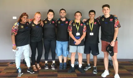 Group of six youth and one adult, all Indigenous, wearing black tops and standing arm in arm on a carpeted floor in front of a blank wall.