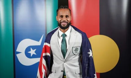 A young man in pale coat and green tie stands with a blue, red and white flag over his shoulders. In the background are two more flags. At left it is green, black and blue with a white symbol at centre. At right is a red and black flag with a yellow disc