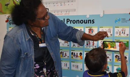 A woman and a young boy stand in front of a poster covered with small images depicting pronouns in language. The young boy holds an iPhone to one of the images to activate the augmented reality. The large poster hangs on a wall along with other posters.