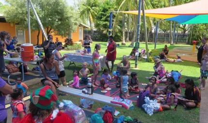 Large group of children and adults sit on grass and at tables under shade, dressed in swimming attire and other causal clothes. In the background are trees, shade covers and a building.