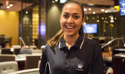 A young Indigenous woman dressed in black polo neck shirt with a Crown insignia stands in a resort lounge featuring lights, booths and chairs.