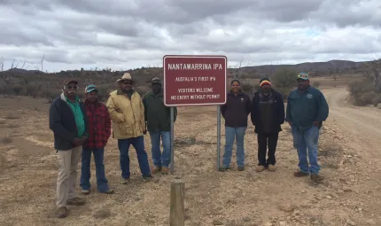 Seven Aboriginal men and women dressed in warm clothing stand next to a red sign just off a dirt road. In the background are bushes, trees and hills. The sign says Nantawarrina IPA – Australia’s first IPA – Visitors welcome – No entry without permit.