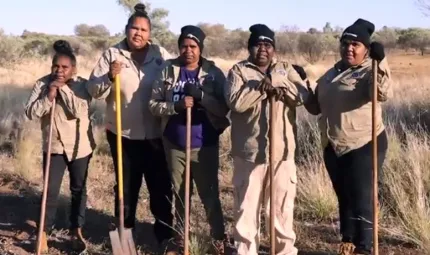 Five women dressed in work wear hold large gardening implements (shovels) and face the camera. In the background is dried grass, bushes and blue sky.