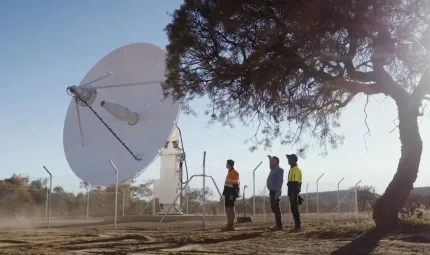 A large and white satellite dish sits on a concrete slab. Three men, two of whom are in work wear look at it. At right is a tree and in the background are hills and vegetation.