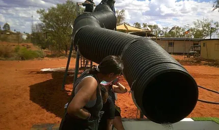 A long and very wide black pipe snakes from a stand downward to a pool. On the stand are several people and at the pool are a few more. In the background is red soil, trees and buildings.