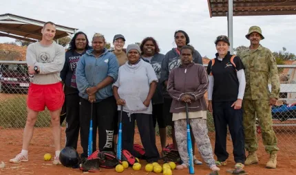 Indigenous women with non-Indigenous army men and women wearing various clothing types and some holding softball bats. There are softballs on the red earth in front of them.