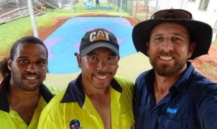 Three men, two in yellow work-wear shirts and one in a blue shirt stand close to camera. In the background is a flat area covered with blue and pink rubber. Surrounding this is bare soil, grass and fences. There are buildings at right and left and behind.