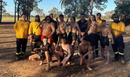 Group of young men dressed in traditional wear and paint stand or kneel on dry ground. With them are men in firefighting wear. In the background are trees, a grassy field and a blue sky.