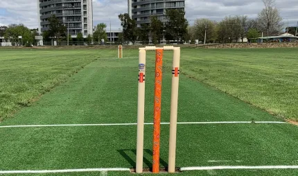 Two sets of cricket stumps and bails stand at each end of a narrow green strip of artificial grass. The two middle stumps are bright orange with Indigenous designs. On both sides of the pitch is real grass and in the background are two tall buildings.