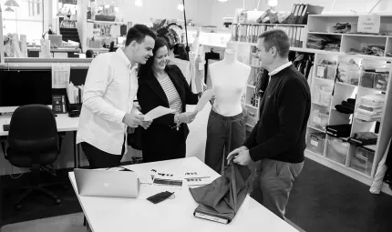 Two men and a woman in casual clothing stand next to a table on which is a laptop, some papers and a roll of material. In the background is a desk and shelves filled with papers, boxes and office supplies.