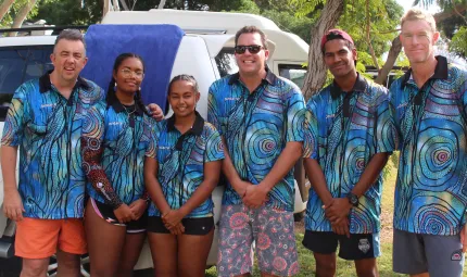 6 adults (2 female and 4 male / 3 Aboriginal) dressed in blue shirts featuring Indigenous designs stand in front of a white car on grass with trees in the background.