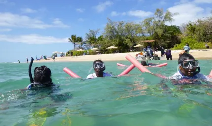 Three Indigenous people wearing snorkles and diving masks, and holding floating noodles swim in seawater. In the background is sand, beach umbrellas, trees and other people.